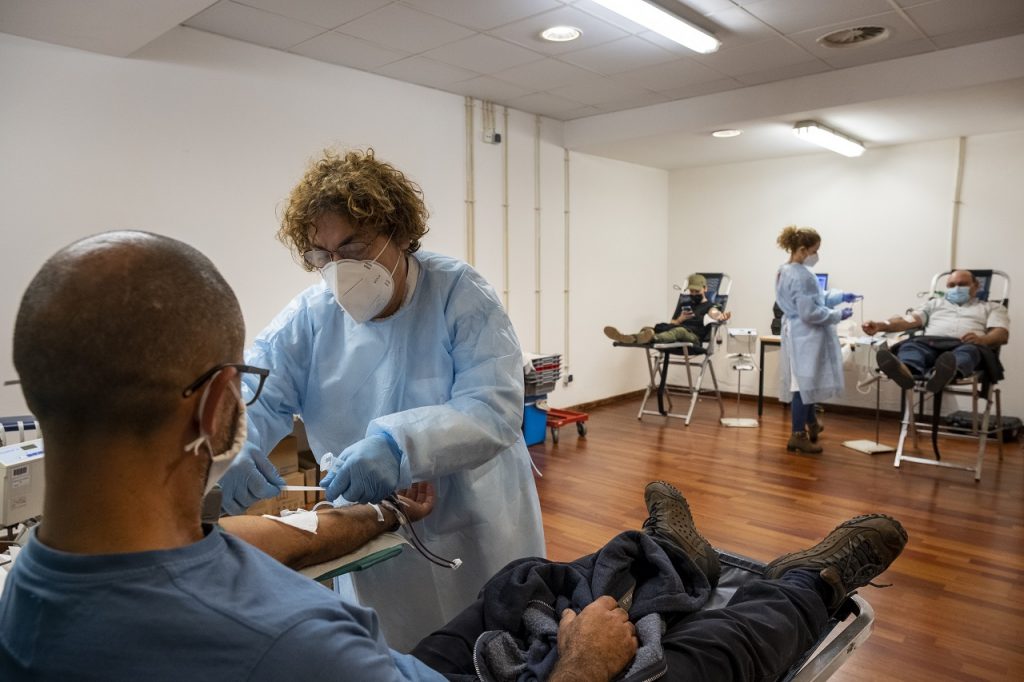 Três pessoas estão sentadas em cadeirões para dar sangue com apoio de duas enfermeiras